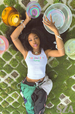 SZA lying on grass with colorful bowls and a drink, wearing a white tank top with text.